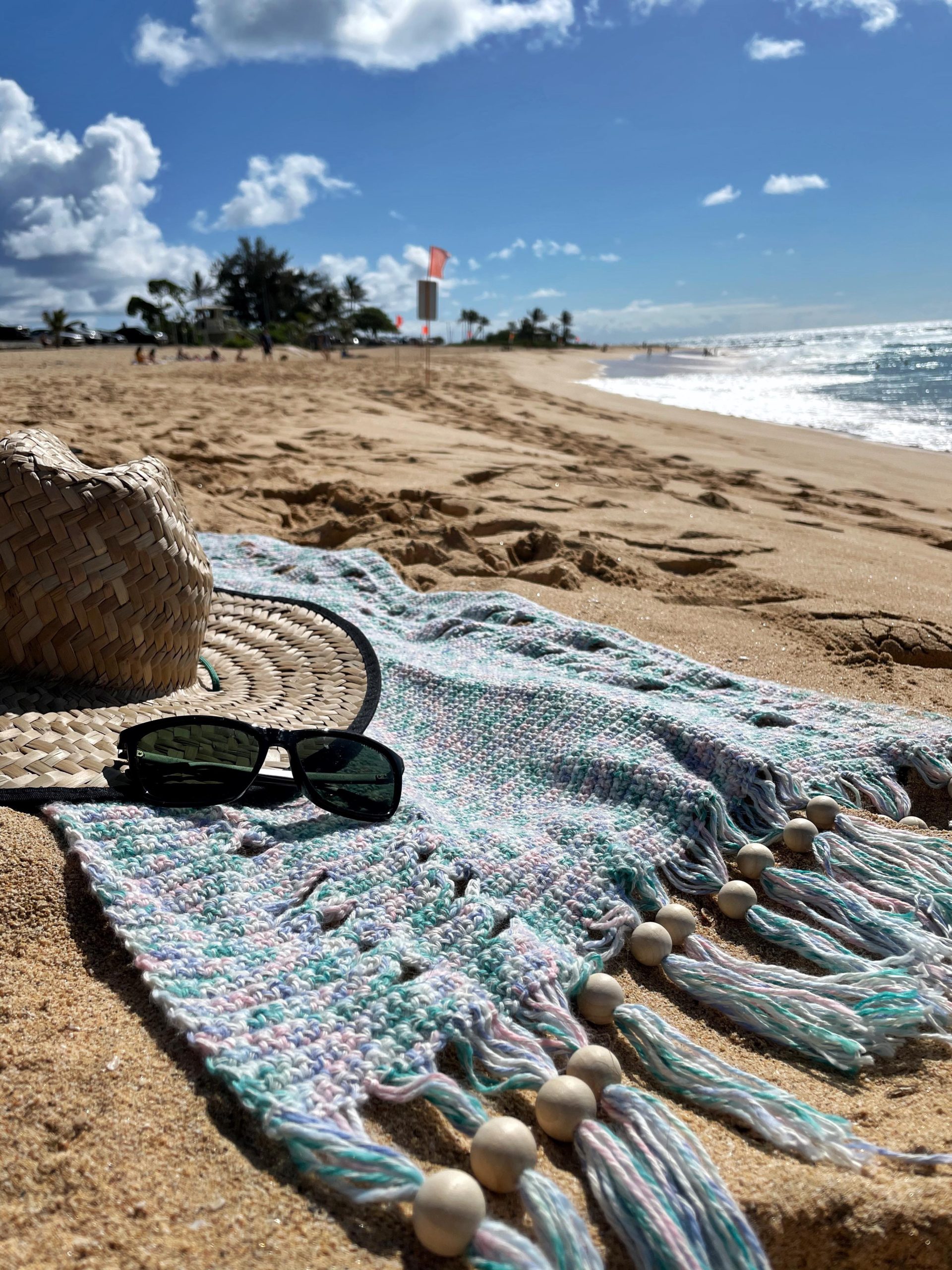 crochet beach towel on a sandy beach with a hat and sunglasses on top of it