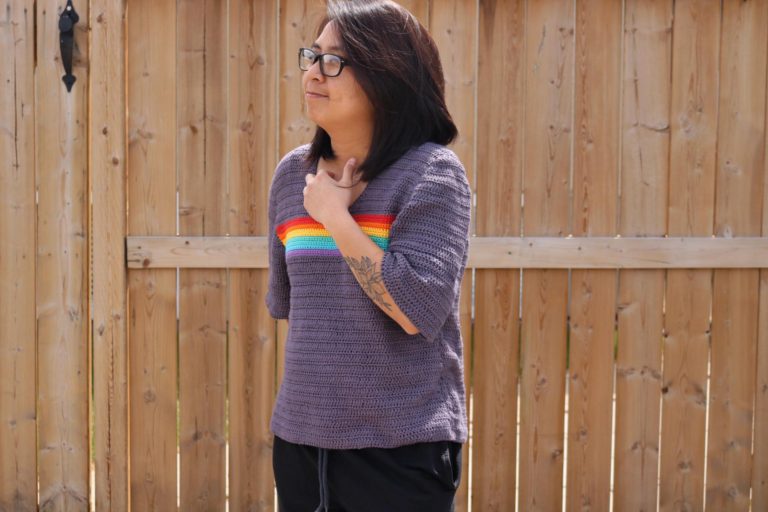 person standing in front of a wooden fence wearing a gray crochet shirt with a band of rainbow stripes across the mid chest area.
