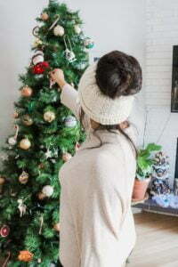 Girl putting on Christmas tree ornaments wearing a white <span style='background-color:none;'>messy bun beanie</span><span style='background-color:none;'> </span>and a cream long sleeved top.